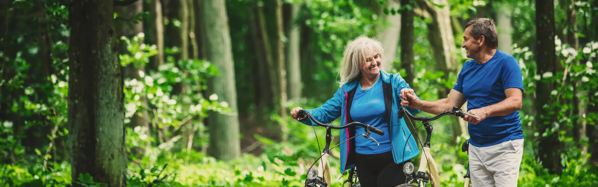 Middle aged white couple stands in the middle of a green forest. Both of them are standing beside bicycles. 