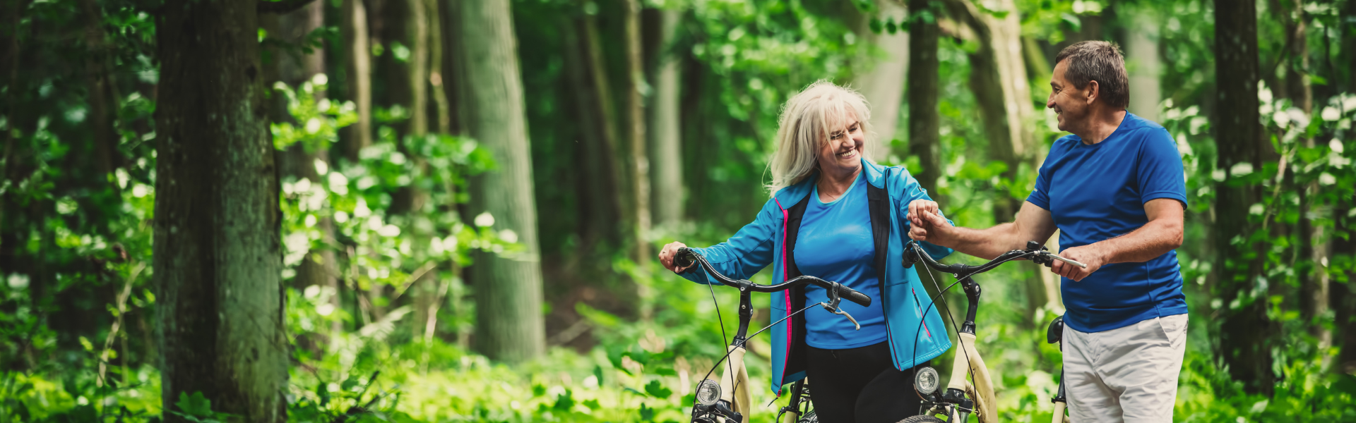 Middle aged white couple stands in the middle of a green forest. Both of them are standing beside bicycles. 